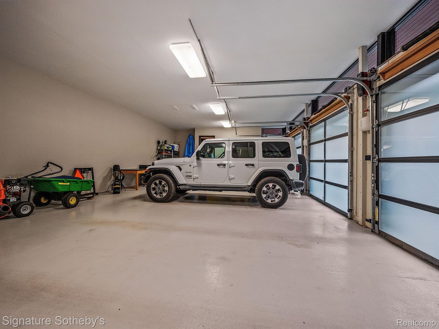 The image showcases a spacious garage featuring a white Jeep Wrangler parked inside. The garage has a modern aesthetic with a glass panel garage door, bright overhead lighting, and a clean, polished floor. Additional equipment like a lawn mower and garden cart are neatly stored along the wall, indicating ample space and functionality.