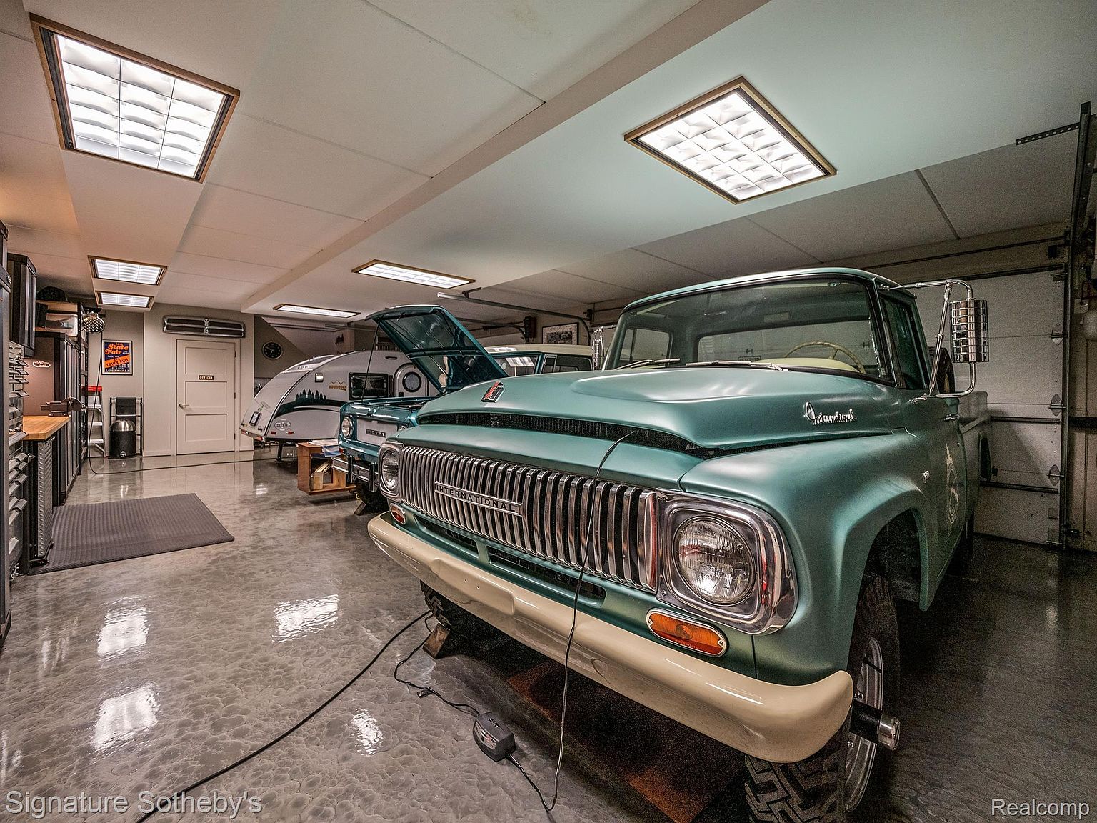 This image showcases a well-maintained garage featuring a classic teal truck as a focal point, complemented by a vintage camper in the background. The garage boasts a clean, polished floor and ample lighting, creating an inviting space for car enthusiasts or hobbyists. The perspective is from a low angle, emphasizing the size and depth of the garage.