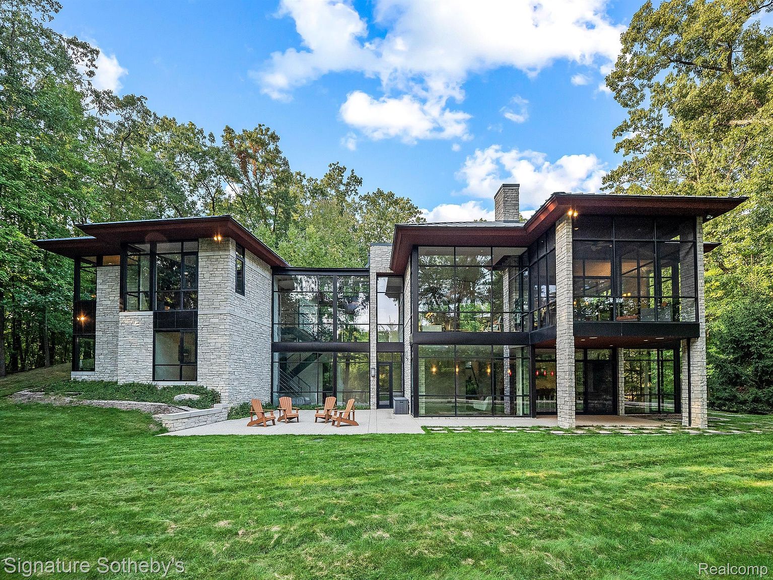 This is a rear view of a modern, luxurious home featuring a stone facade and expansive glass windows that offer abundant natural light. The property boasts a well-manicured lawn and a patio area with outdoor seating, perfect for entertaining. The architectural design emphasizes clean lines and a seamless integration with the surrounding wooded landscape.