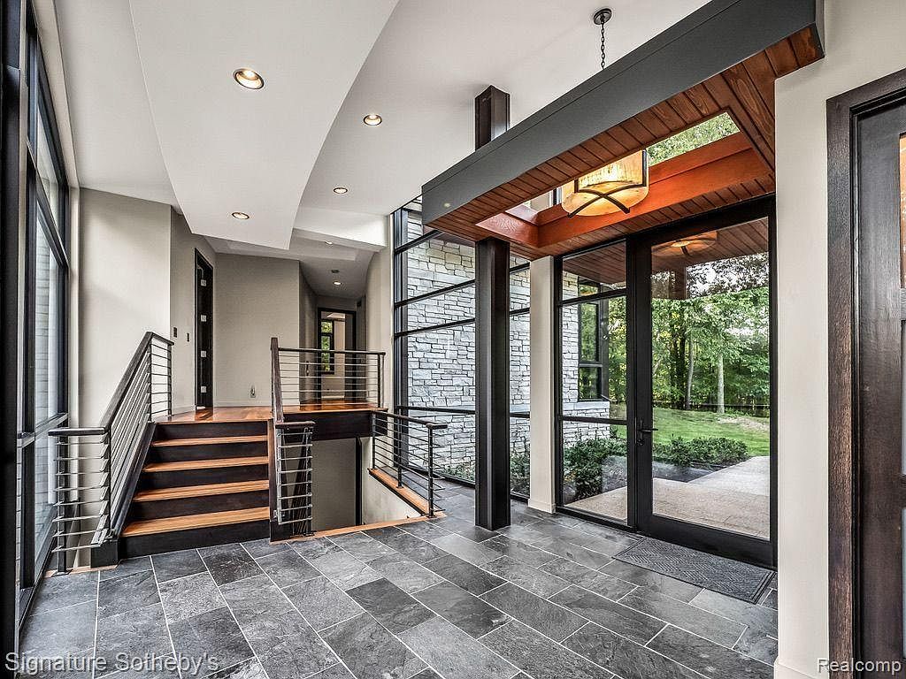 This interior shot showcases a modern entryway featuring dark gray slate flooring and a unique split-level staircase with wood treads and stainless steel railings. A striking architectural element with wood and dark metal framing houses a contemporary light fixture and provides a view to the exterior. The space is well-lit and exudes a sophisticated, contemporary aesthetic.