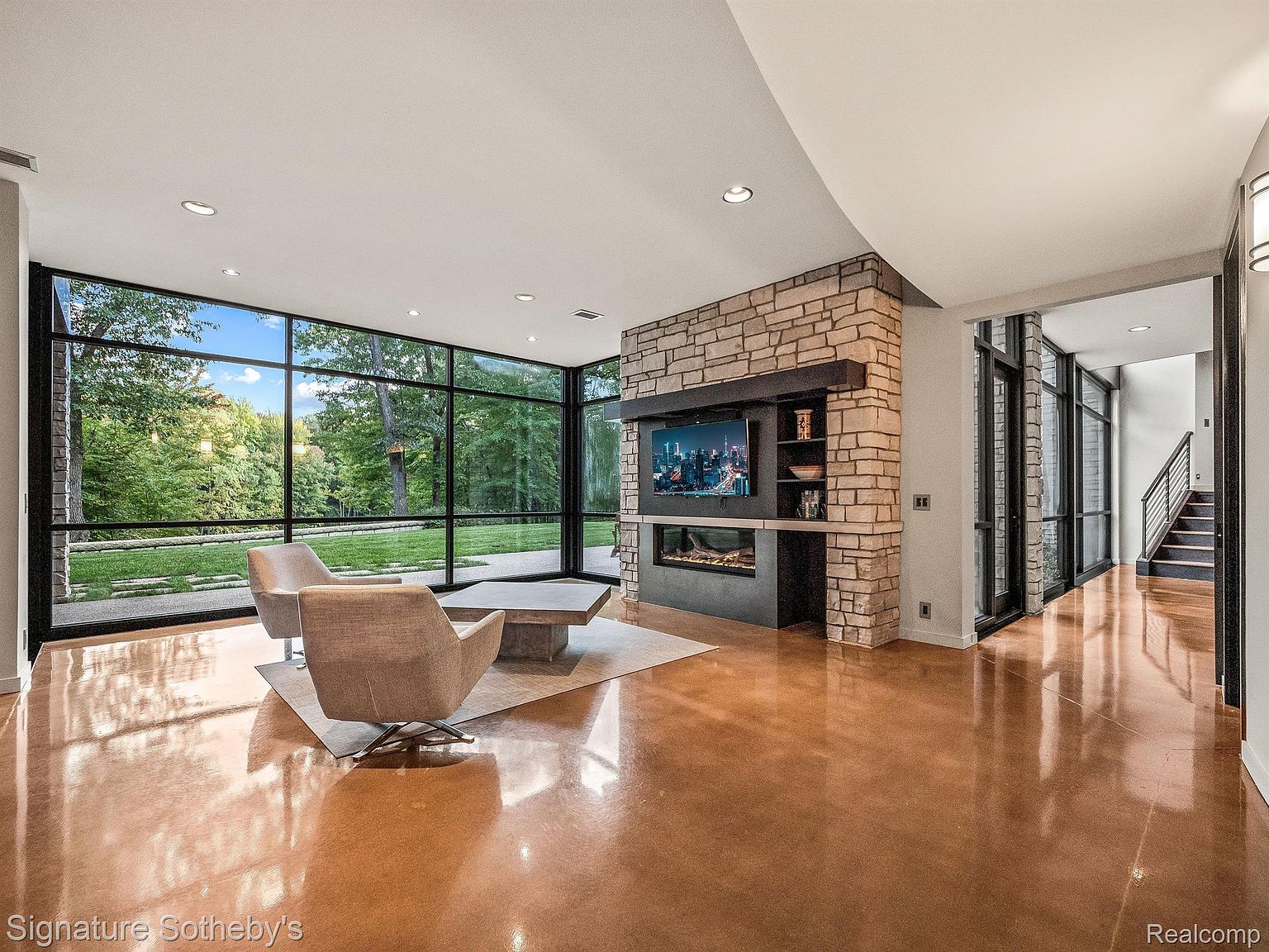 This is a modern living room featuring a large window wall with views of a lush green landscape. The room includes a stone fireplace with a built-in TV, two armchairs, and a coffee table on a light-colored rug. The polished concrete floors reflect light, enhancing the spacious and airy feel of the room.