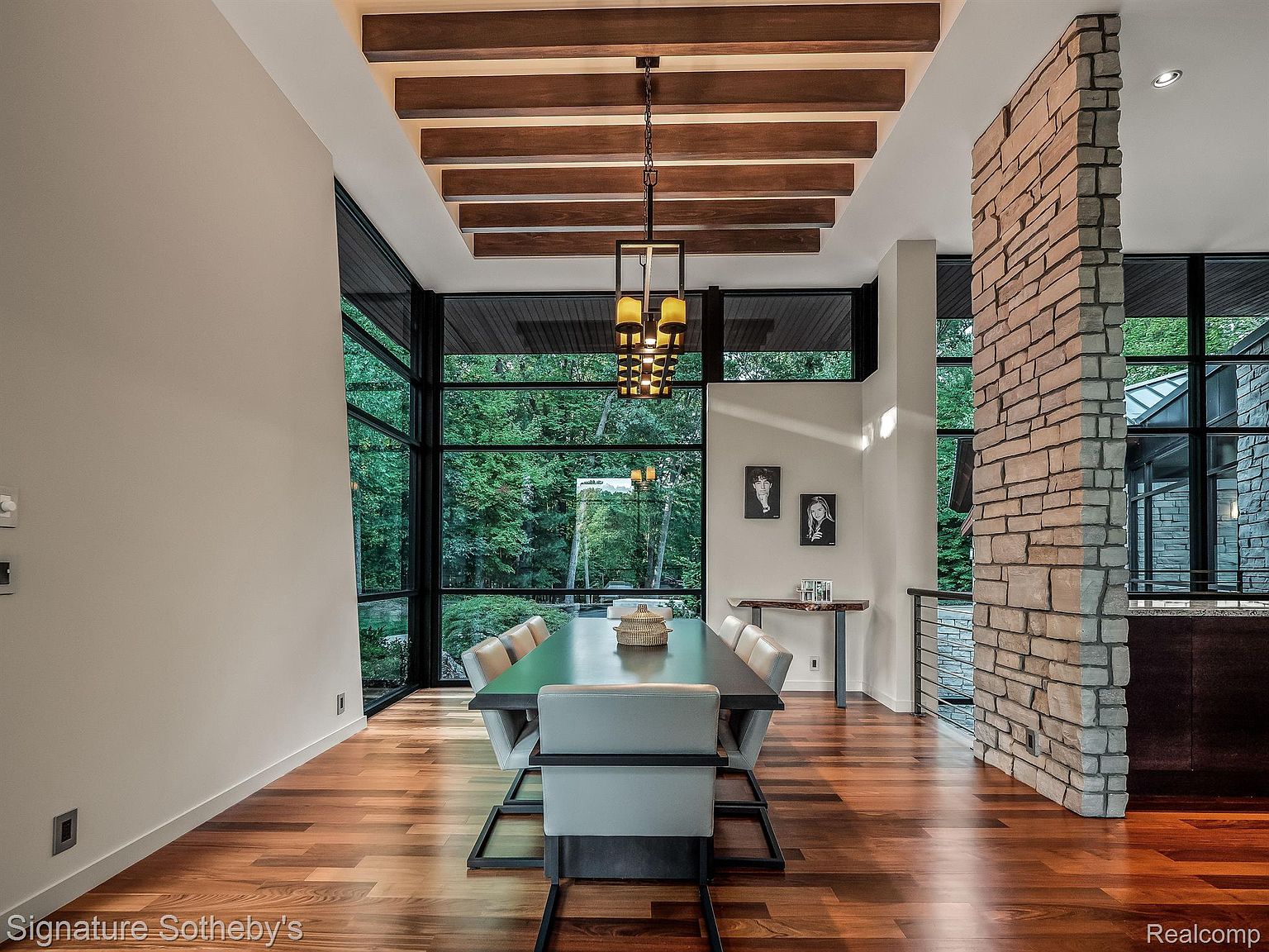 This is an interior shot of a dining room featuring a long, dark wood table surrounded by modern chairs with light upholstery. A striking light fixture hangs above the table, and a large window offers a view of the surrounding greenery. A stone accent wall adds texture and visual interest to the space, complemented by hardwood flooring throughout.