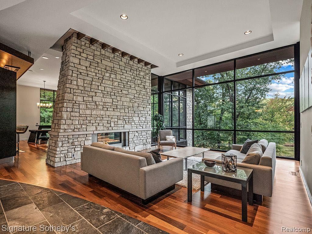 This is a spacious living room featuring a stone fireplace that acts as a central architectural element. Large windows offer expansive views of the surrounding greenery, creating a seamless connection with nature. The room is furnished with modern sofas and tables, complemented by hardwood flooring, creating a warm and inviting atmosphere.