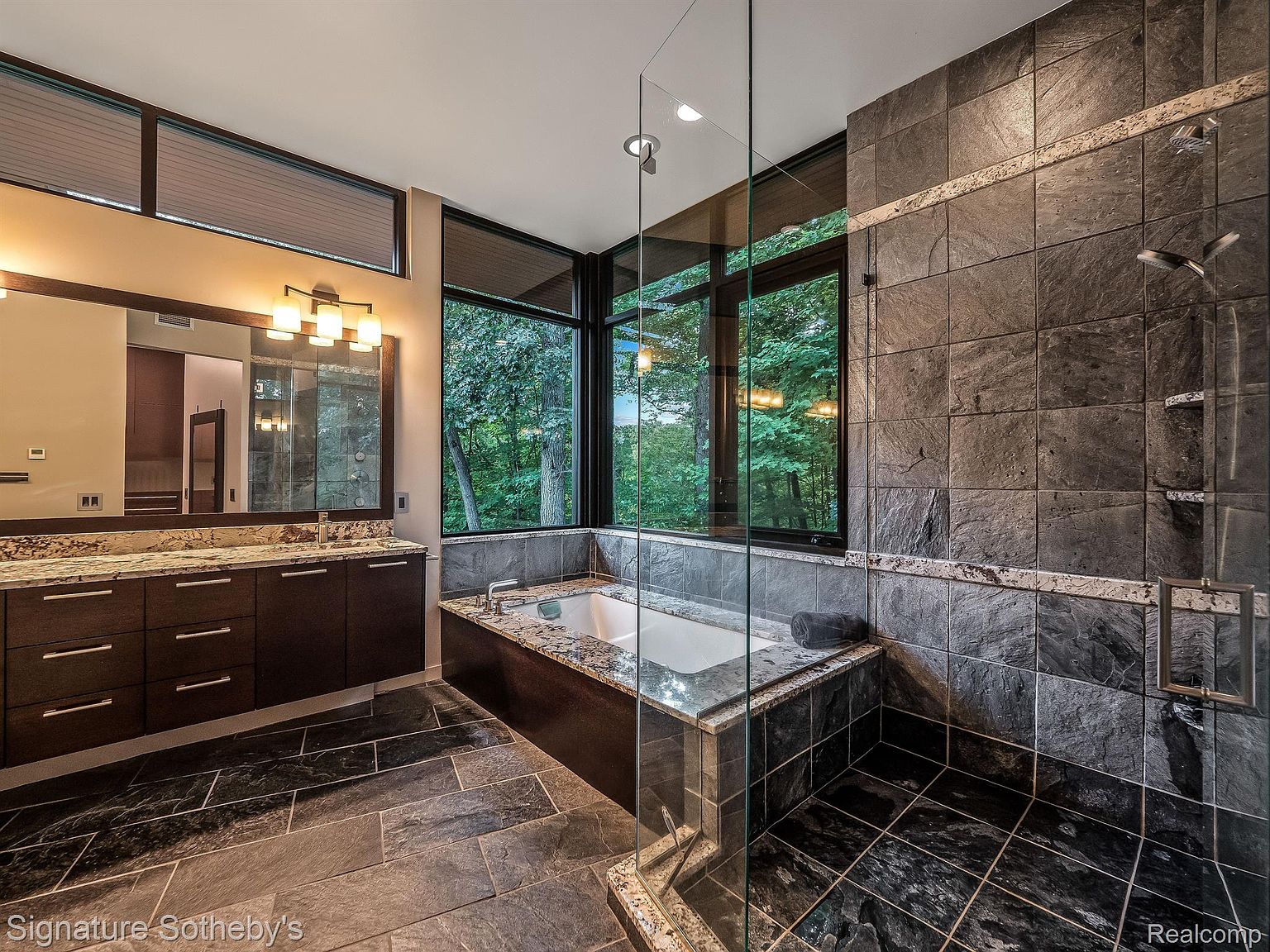 This is a luxurious primary bathroom featuring dark stone tile flooring and wall accents. A large soaking tub is positioned beneath a window offering a view of the surrounding greenery, while a glass-enclosed shower is located adjacent to the tub. The vanity area includes a double sink with a granite countertop and dark wood cabinetry, creating a spa-like atmosphere.