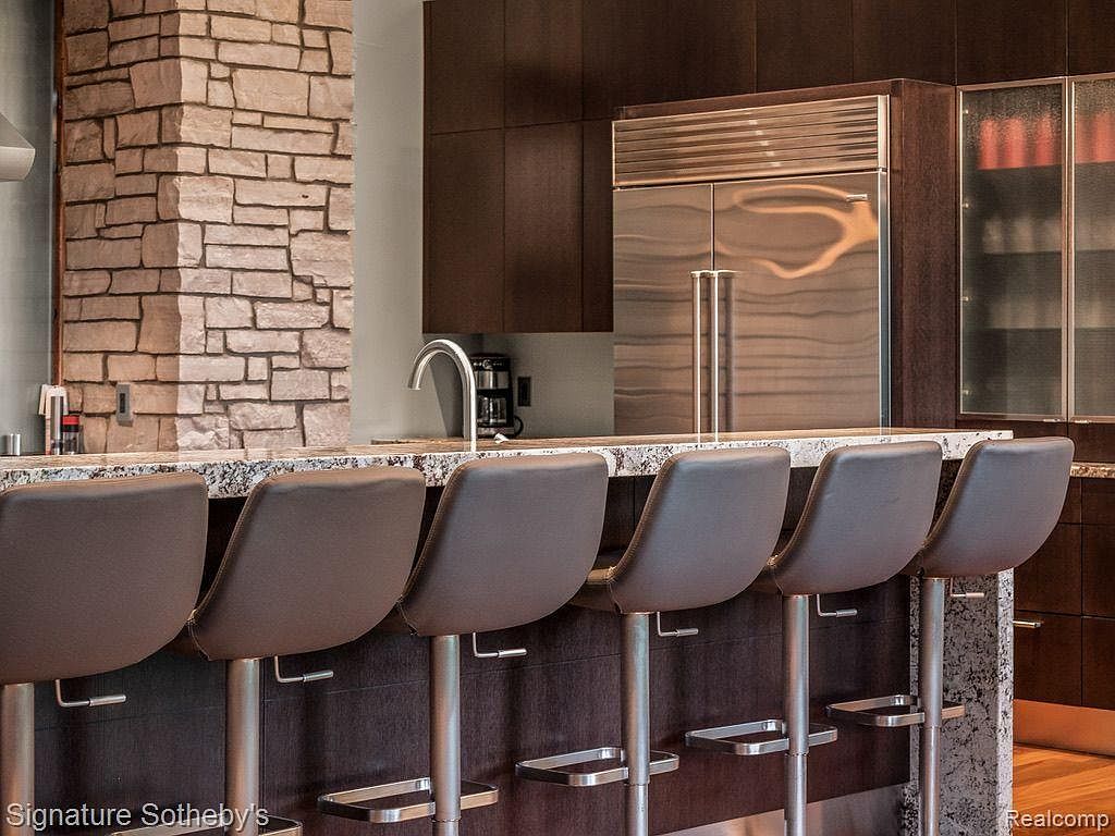 This is a view of a modern kitchen featuring a granite countertop with six bar stools. The kitchen includes dark wood cabinetry, a stainless steel refrigerator, and a stone accent wall. The overall impression is one of luxury and sophistication.