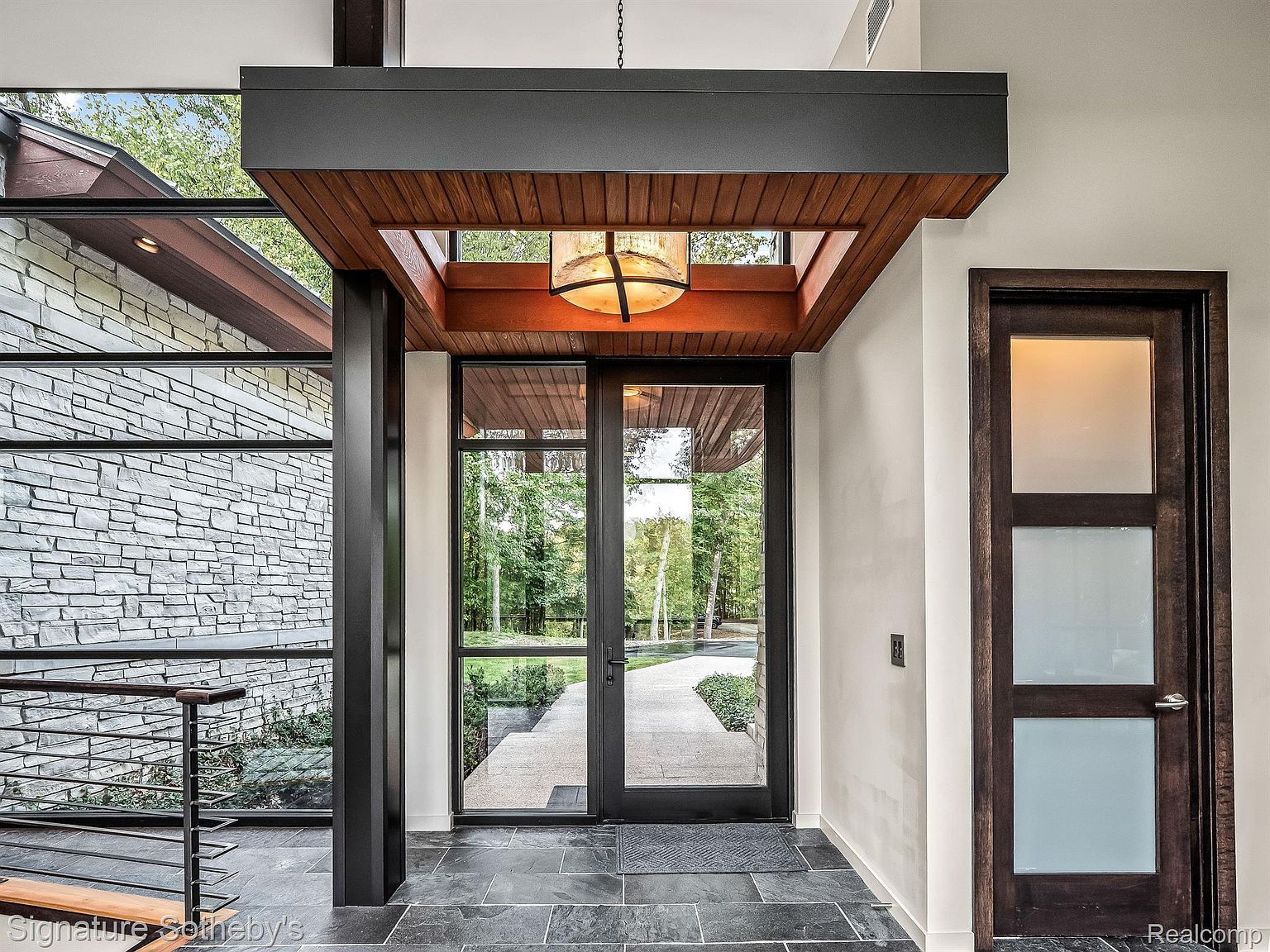 This image showcases a modern home's entryway, featuring a striking combination of stone and glass exterior walls. The entrance is defined by a dark-framed glass door under a wood-paneled overhang with a unique light fixture. The interior flooring is dark tile, and a frosted glass door is visible to the right, adding to the contemporary aesthetic.