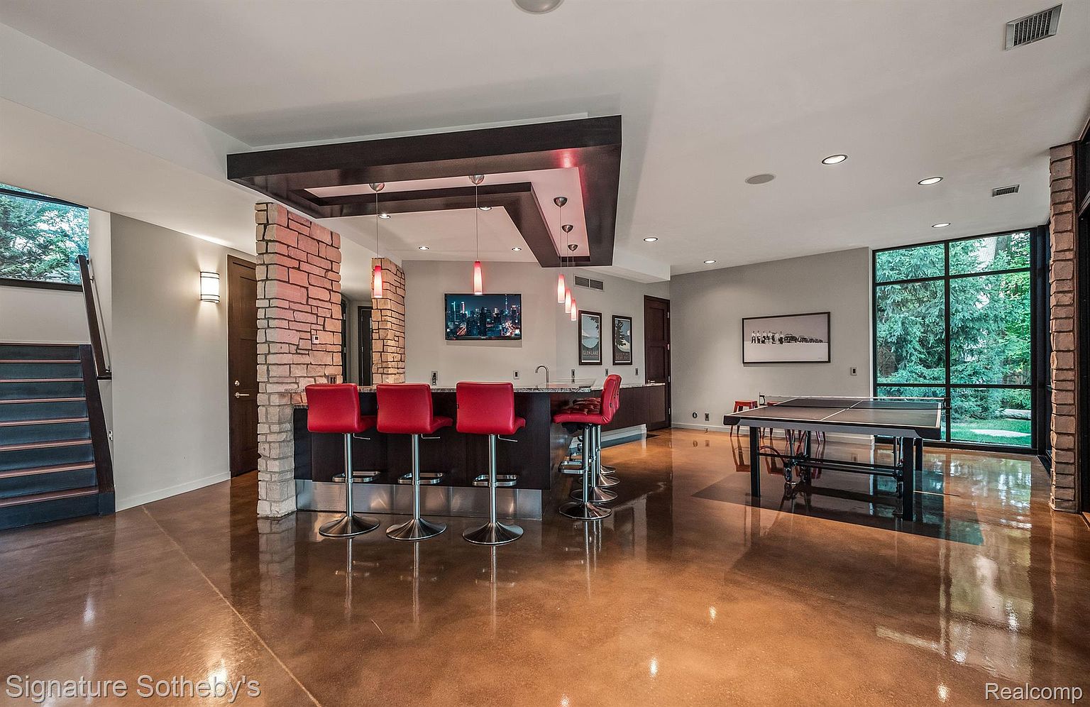 This is a spacious basement area featuring a bar with red bar stools, a brick accent wall, and a ping pong table. The polished concrete floors reflect the ambient light, creating a warm and inviting atmosphere. Large windows provide natural light and a view of the outdoors.