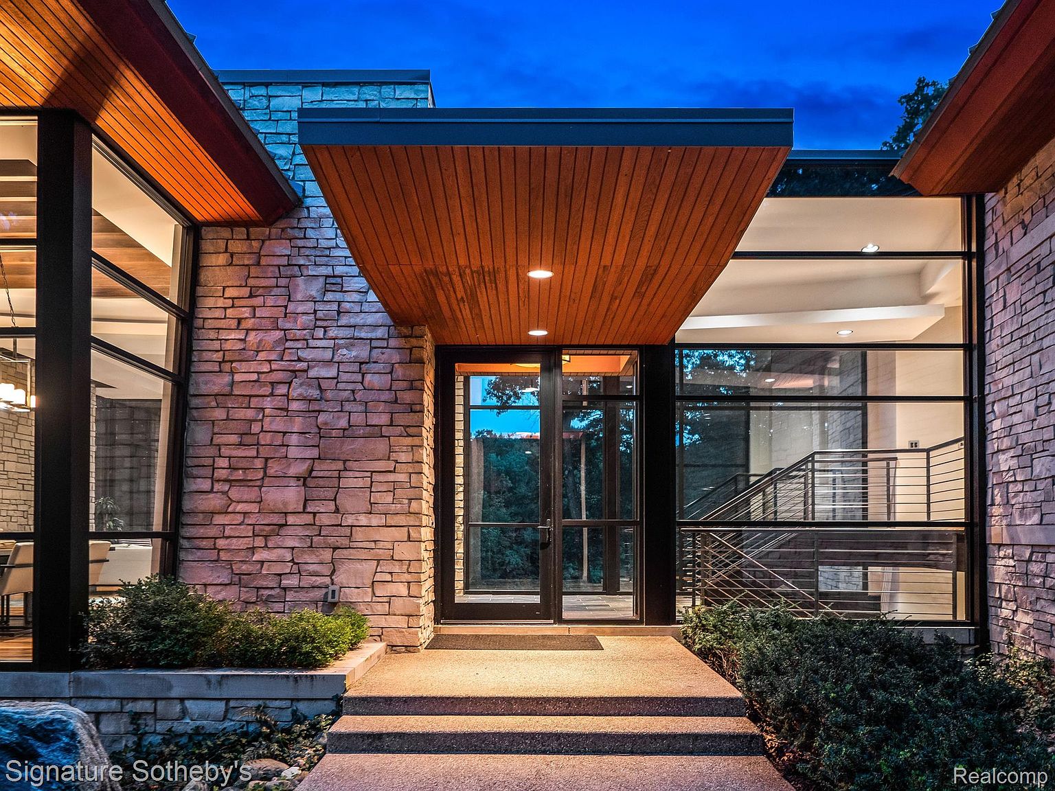 This image showcases the grand entryway of a modern home, featuring a stone facade, a striking wood-paneled overhang, and large windows that offer a glimpse into the interior. The entrance is accessed by a set of concrete steps, flanked by manicured greenery, creating an inviting and sophisticated first impression. The perspective is from the front, emphasizing the architectural details and the welcoming nature of the home.