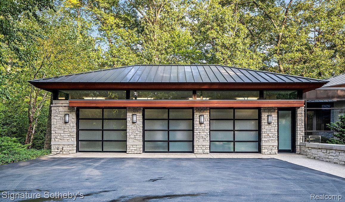 This image showcases a modern garage exterior with three frosted glass doors framed by stone pillars and black trim. A low-pitched roof with dark metal panels and wood trim overhangs the structure, while the driveway is paved with asphalt. The garage is surrounded by lush green trees, creating a sense of privacy and seclusion.