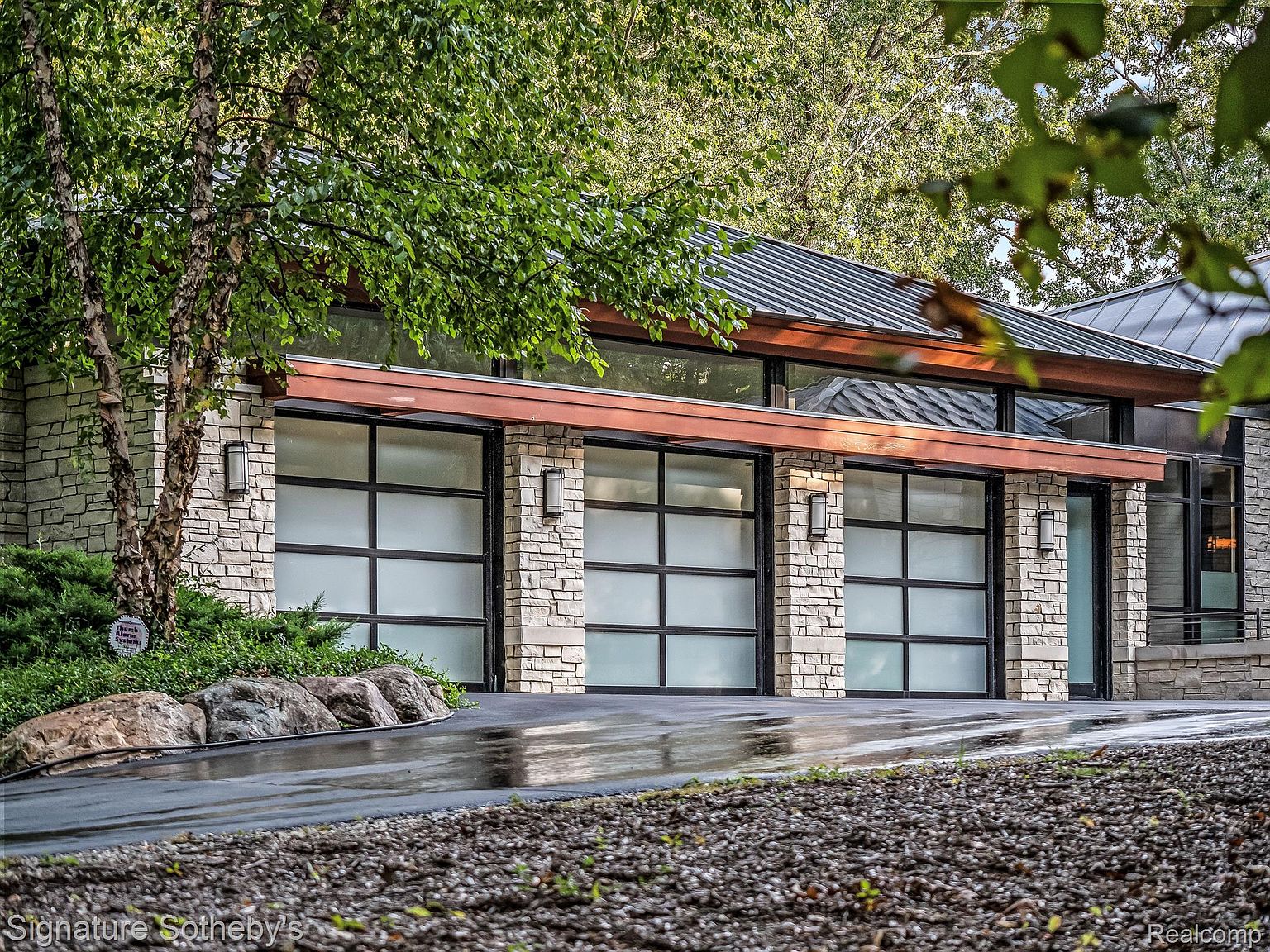 This image showcases a modern garage exterior featuring three frosted glass garage doors with black frames, set within a light-colored stone facade. The structure is topped with a metal roof and accented by copper-colored trim. The driveway is wet, reflecting the surrounding greenery and architecture, creating a sleek and contemporary aesthetic.