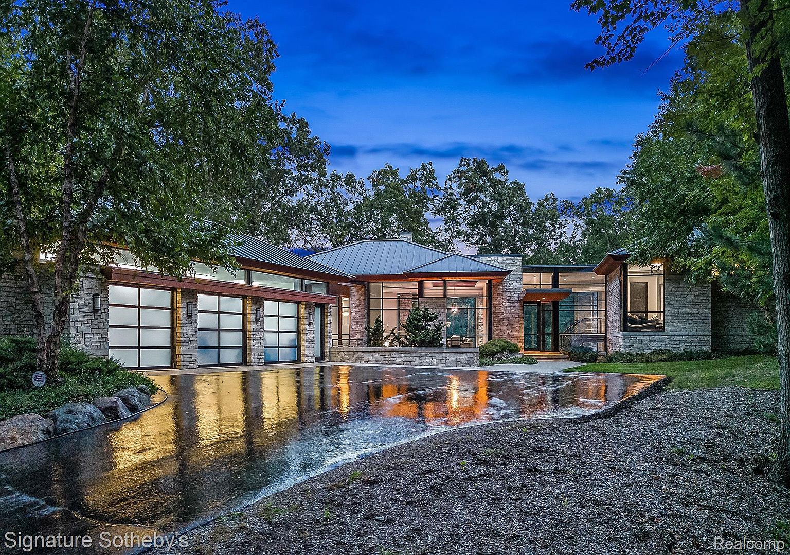 This is a front exterior view of a modern, single-story home with a stone facade and a metal roof. The house features large windows and multiple garage doors, suggesting ample parking space. The wet driveway reflects the ambient light, creating a sleek and inviting appearance, while mature trees frame the property, adding to its privacy and curb appeal.