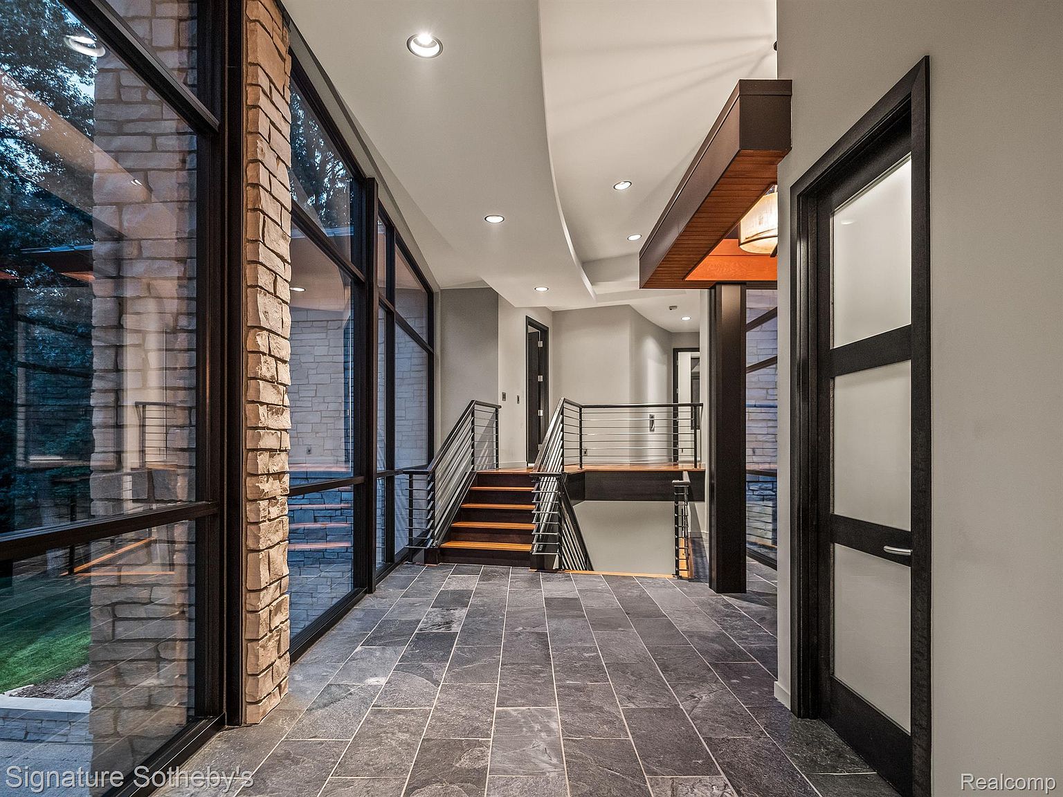 This interior shot showcases a modern hallway with striking architectural details, including large windows framed in black, allowing natural light to flood the space and highlight the stone accent walls. A staircase with sleek metal railings leads to another level, and the dark tile flooring adds a touch of sophistication, creating a visually appealing and well-lit transition space within the home.