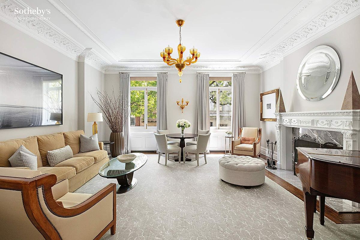 This elegant living room features a sophisticated design with a neutral color palette, accented by a striking gold chandelier. The room is furnished with a comfortable sofa, armchairs, and a round dining table set near the windows, creating a versatile space for both relaxation and entertaining. Architectural details like the ornate ceiling moldings and fireplace add to the room's luxurious appeal, while the large windows provide ample natural light.