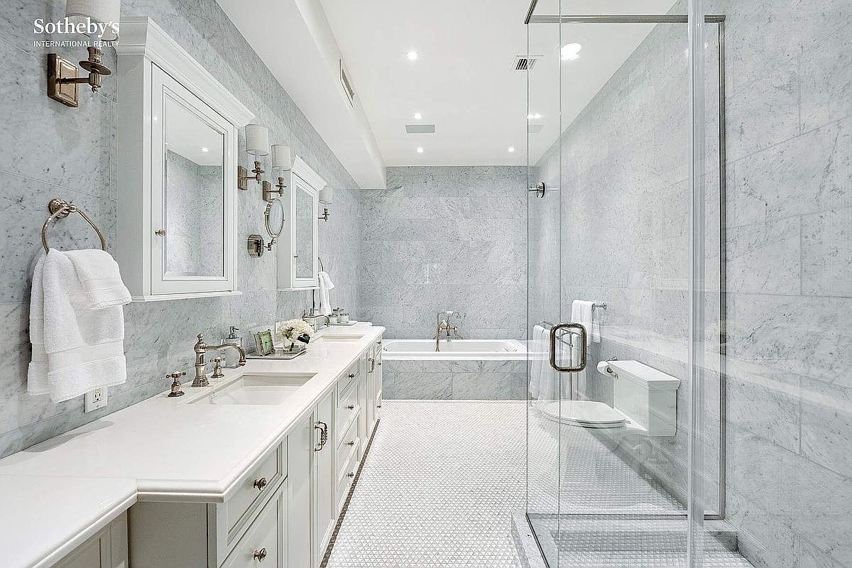 This is a luxurious primary bathroom featuring marble walls and flooring. The bathroom includes a long vanity with dual sinks, white cabinetry, and elegant fixtures. A glass-enclosed shower and a soaking tub are also visible, creating a spa-like atmosphere.
