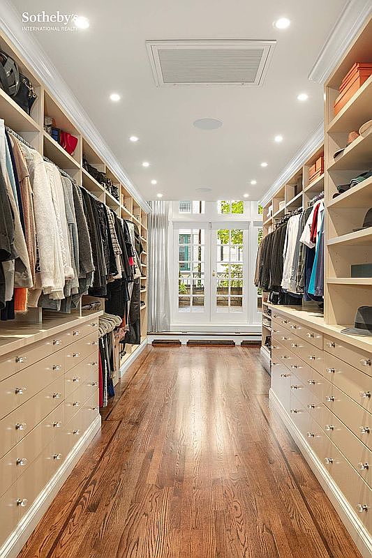 This is a well-organized walk-in closet featuring custom shelving and drawer units on both sides, maximizing storage space. The closet has a hardwood floor and is brightly lit with recessed lighting, creating a luxurious and functional space. A set of double doors at the end of the closet leads to an outdoor area, providing natural light and ventilation.