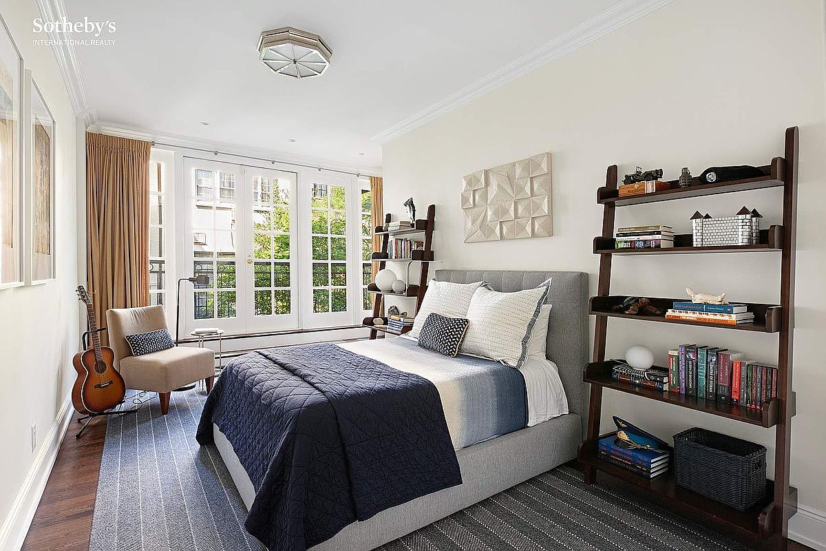 This is a well-lit bedroom featuring a gray upholstered bed with a navy blue quilt, complemented by a ladder-style bookshelf on the right and a smaller one on the left. French doors lead to an outdoor area, enhancing the room's appeal. The room is decorated with modern art and stylish accessories, creating a comfortable and inviting atmosphere.