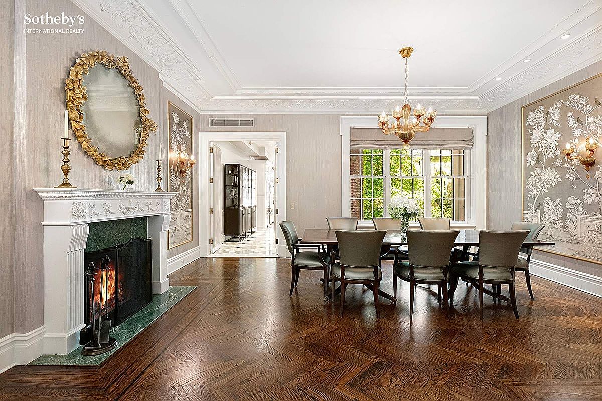This is an elegant dining room featuring a herringbone wood floor, a fireplace with a green marble hearth, and a large oval mirror above the mantel. A dining table with upholstered chairs sits beneath a chandelier, and a large window provides natural light. The room is decorated in neutral tones with ornate moldings and decorative wall panels, creating a sophisticated and inviting atmosphere.