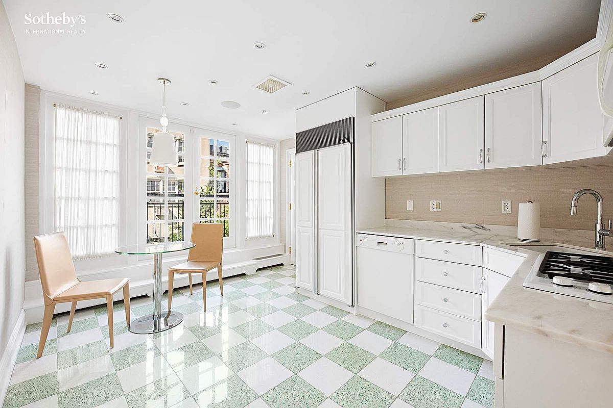 This is a bright and airy kitchen featuring white cabinetry, stainless steel appliances, and a unique checkered floor pattern. The kitchen includes a dining area with a glass-top table and two chairs, positioned near large windows that provide ample natural light. The overall impression is clean, modern, and inviting, with a touch of retro charm from the flooring.