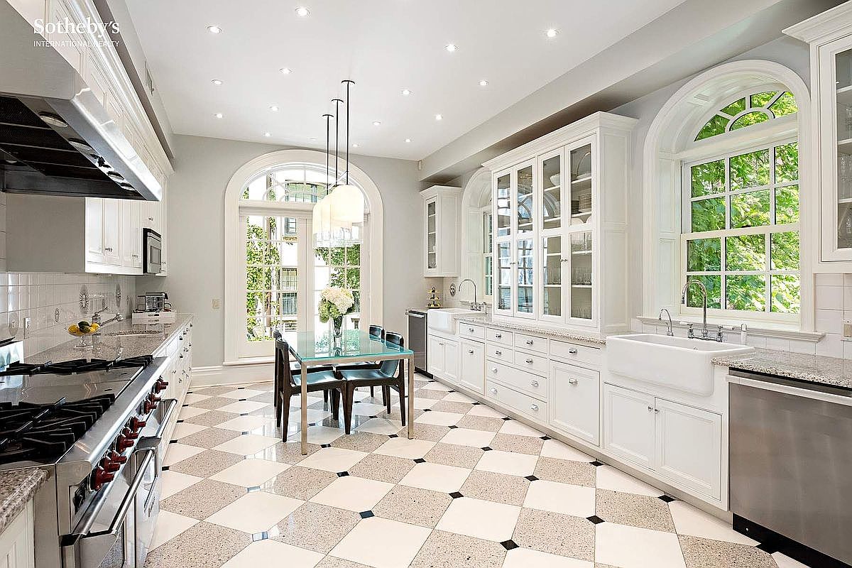 This is a bright and spacious kitchen featuring a checkered tile floor, white cabinetry, and stainless steel appliances. The kitchen is well-lit with recessed lighting and natural light from large arched windows and a door leading to the outside. A glass-topped dining table with chairs sits in the center of the room, creating an inviting space for meals.