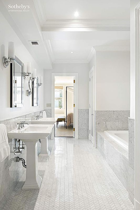 This is a bright and luxurious primary bathroom featuring a long vanity with dual pedestal sinks, each with its own mirror and sconce lighting. The floor is tiled in a herringbone pattern, and the walls are partially covered in marble, adding to the elegant aesthetic. A doorway leads to another room, suggesting a spacious layout.