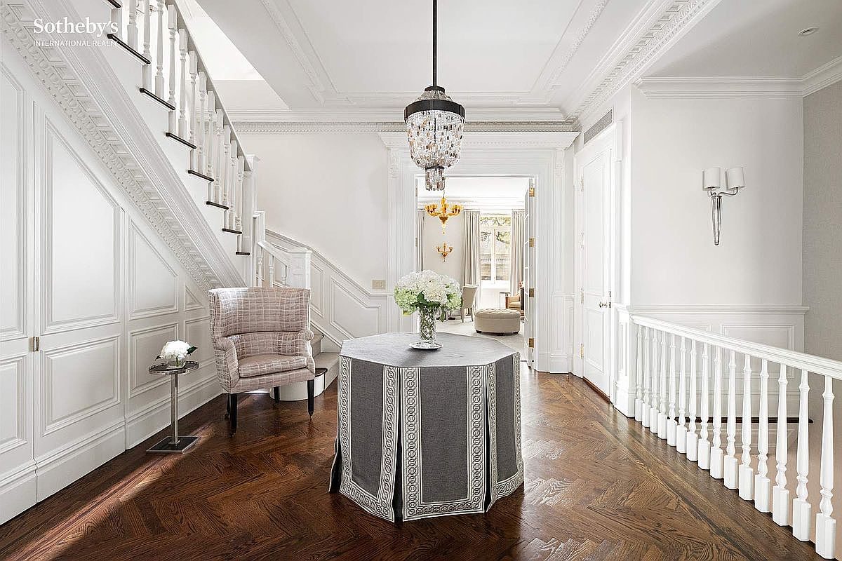 This is an interior shot of a grand hallway featuring a staircase, elegant wainscoting, and dark hardwood floors laid in a herringbone pattern. A round table with a draped cover sits in the center of the hallway, adorned with a floral arrangement, while a crystal chandelier hangs above. The space is bright and airy, with white walls and trim, creating a luxurious and inviting atmosphere.