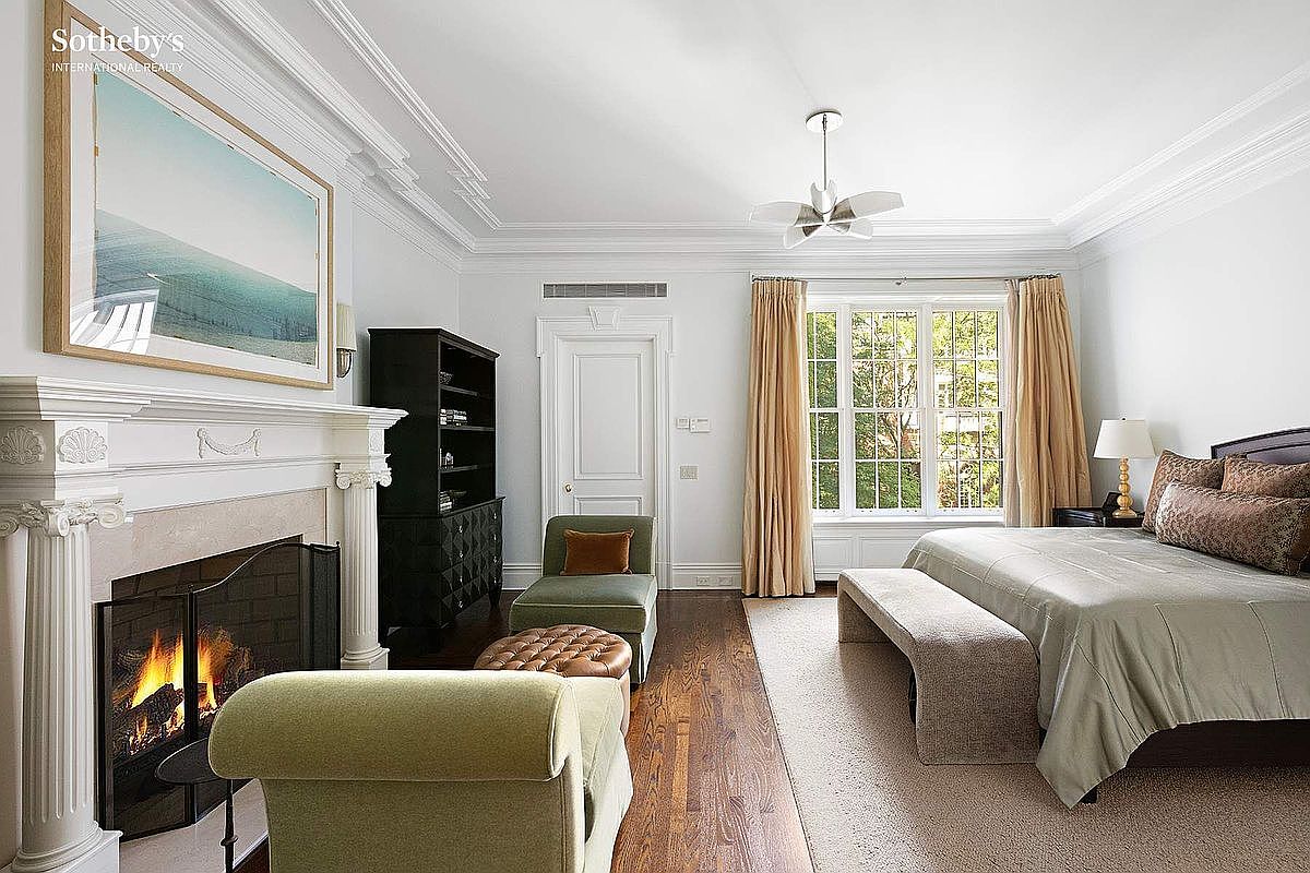 This is a primary bedroom featuring a fireplace with a white mantle, a black bookcase, and a seating area with a chaise lounge. The room has a large window with tan curtains, a bed with a bench at the foot, and hardwood floors with an area rug. The overall style is elegant and comfortable.