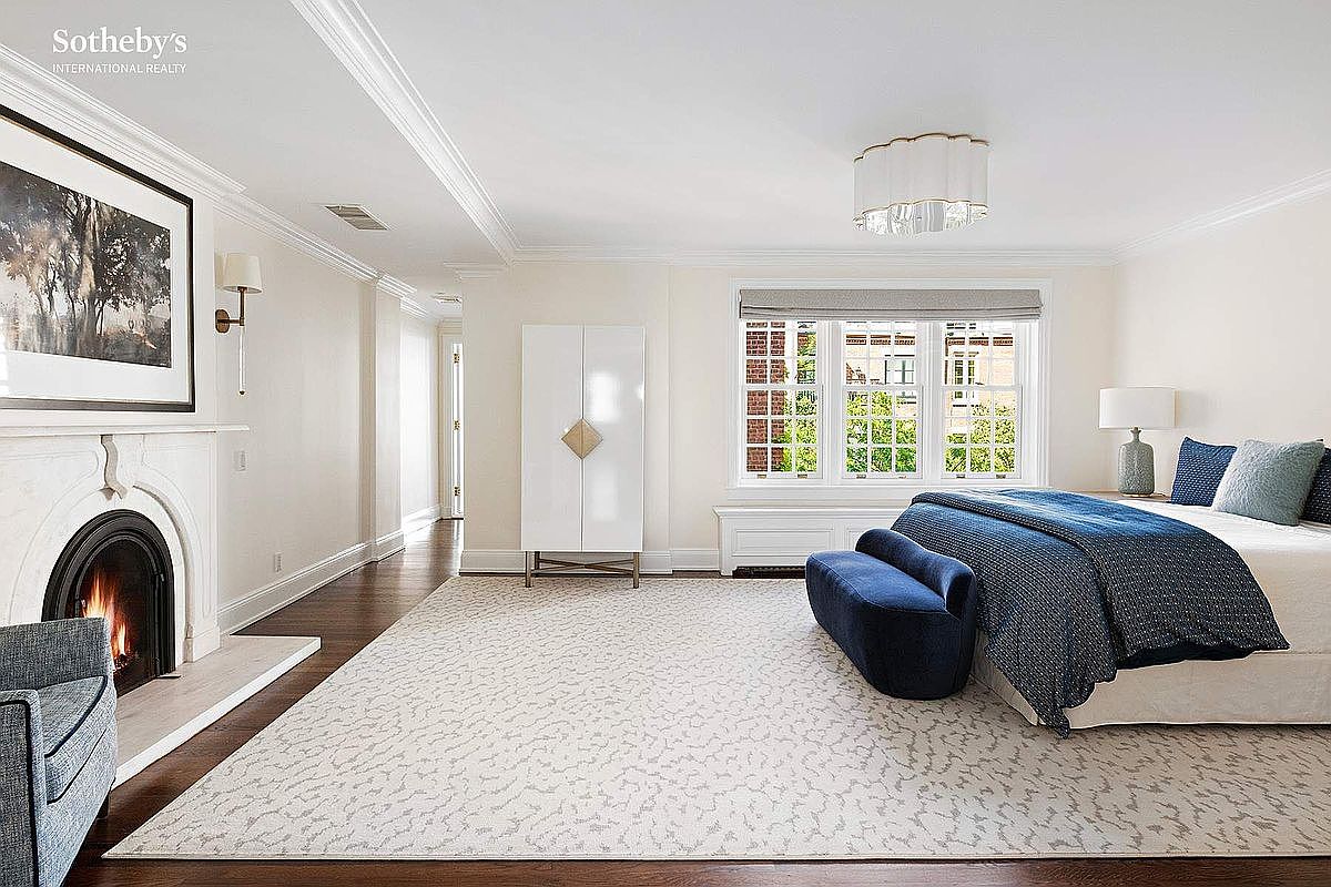 This is a spacious primary bedroom featuring a fireplace with a marble surround, a large window with a view, and a king-size bed with blue accents. The room is decorated in a neutral color palette with hardwood floors and a patterned area rug. A white armoire provides storage, and the overall impression is elegant and serene.