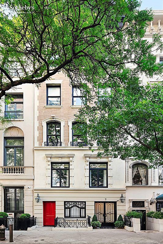 This is a front view of a classic townhouse featuring a red front door and black iron details. The facade is a mix of light-colored stone and brick, with symmetrically arranged windows. Mature trees partially frame the building, adding a touch of greenery and privacy.