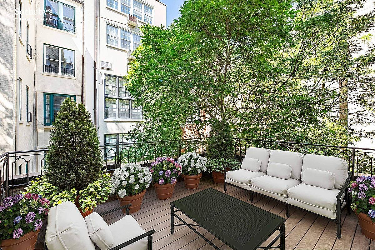 This image showcases a charming outdoor balcony or deck area, furnished with a comfortable white sofa set and a dark metal coffee table. The space is adorned with potted plants, including hydrangeas, creating a lush and inviting atmosphere. The black metal railing adds a touch of elegance, while the backdrop features a building facade and mature trees, enhancing the sense of privacy and tranquility.