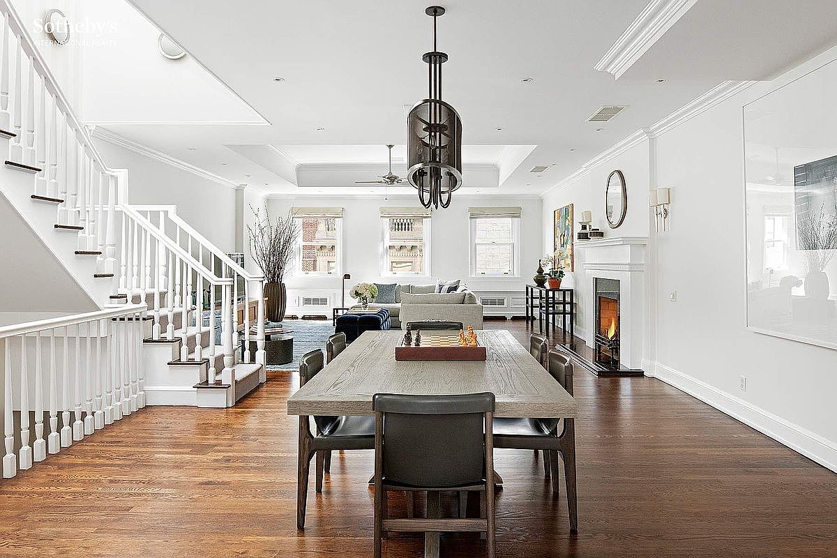 This interior shot showcases a well-lit dining room with hardwood floors and white walls. A large wooden dining table with leather chairs sits prominently in the center, illuminated by a modern chandelier. The room also features a fireplace, staircase, and glimpses of a living area, creating a spacious and inviting atmosphere.