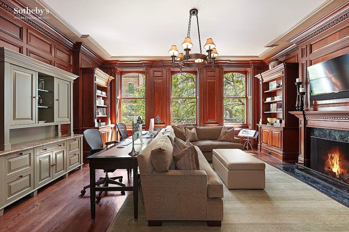 This is a luxurious home office featuring rich wood paneling, built-in bookshelves, and a fireplace with a television mounted above. A large desk and comfortable seating area create a functional and inviting workspace. The room is well-lit by natural light from the windows, offering a view of the outdoors.