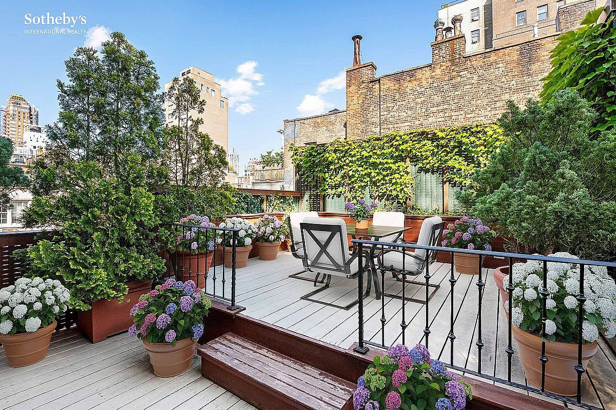 This image showcases a beautifully decorated rooftop patio or deck, perfect for outdoor entertaining. The space features a dining table with chairs, surrounded by lush greenery and potted flowers, creating a serene and private atmosphere. A wrought iron railing adds a touch of elegance, while the wooden deck provides a warm and inviting feel.