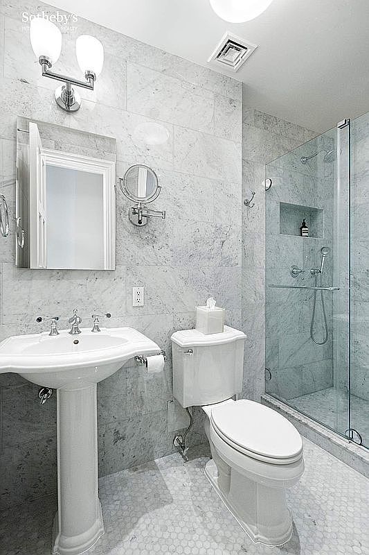 This is a well-lit bathroom featuring marble walls and flooring, giving it a luxurious and clean appearance. The bathroom includes a pedestal sink, a toilet with a tissue holder, a mirrored medicine cabinet, and a glass-enclosed shower. The overall impression is modern and elegant.