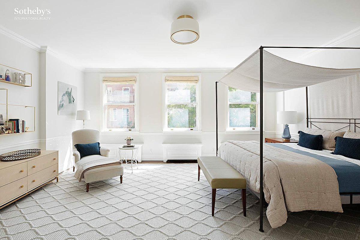 This is a bright and airy primary bedroom featuring a four-poster bed with a light canopy, complemented by a neutral color palette with blue accents. The room includes a comfortable armchair, a dresser with decorative shelving, and a bench at the foot of the bed, all set on a patterned rug. Natural light floods the space through multiple windows, creating a serene and inviting atmosphere.