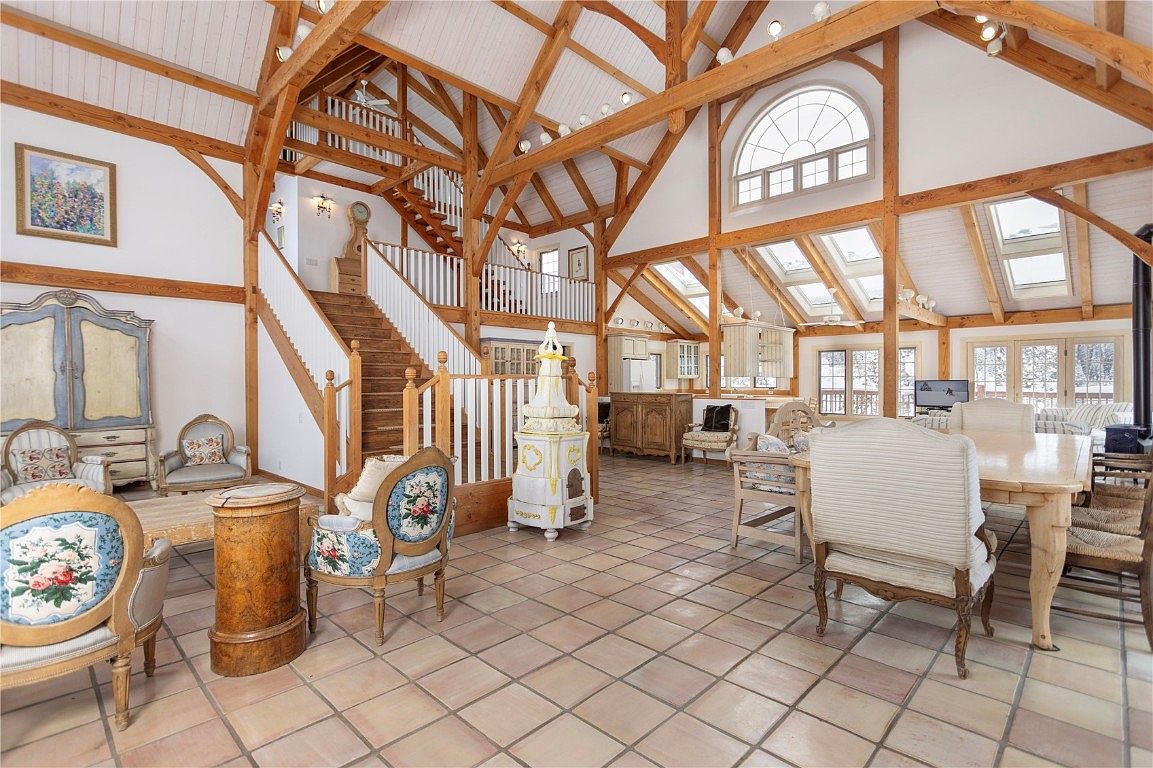 This is a spacious living room with a high vaulted ceiling featuring exposed wooden beams. The room is furnished with antique-style chairs and tables, and a staircase leads to an upper level. Natural light floods the space through large windows and skylights, creating a warm and inviting atmosphere.