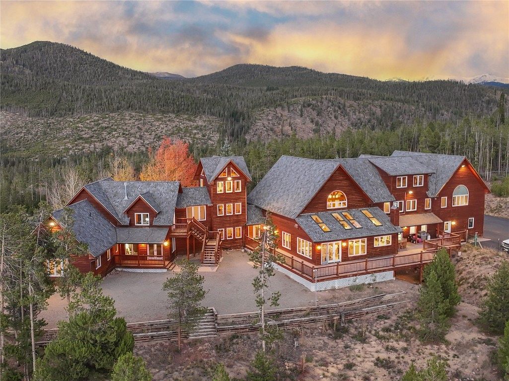 This aerial view showcases a sprawling, multi-sectioned log cabin-style home nestled in a mountainous, wooded landscape. The property features a gray roof, warm wood siding, and multiple decks and balconies, blending seamlessly with its natural surroundings. The overall impression is one of rustic luxury and secluded tranquility.