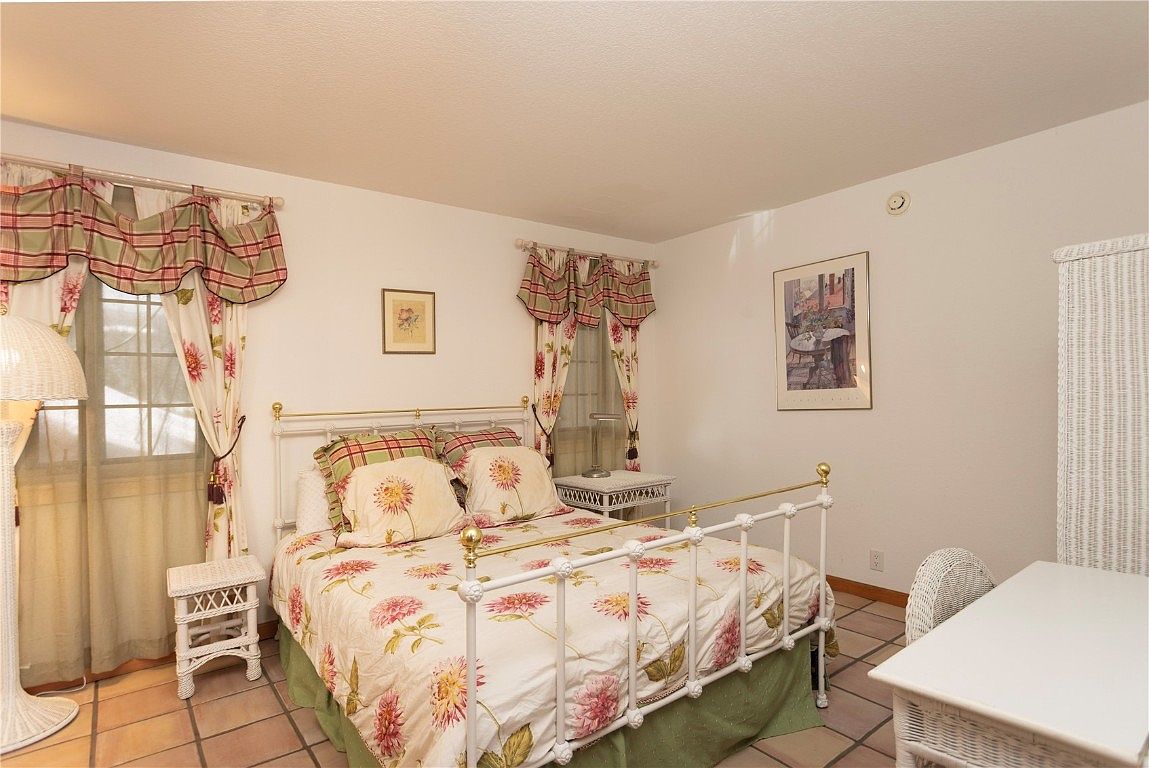 This is a cozy bedroom featuring a white iron bed frame with floral bedding, complemented by matching curtains and valances. The room is decorated with wicker furniture, including a side table and desk, creating a charming and inviting atmosphere. The terracotta tile flooring adds warmth to the space.