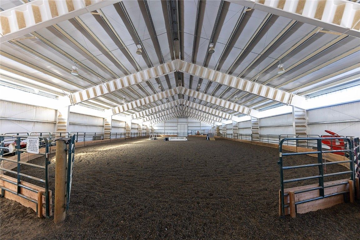 The image showcases a large indoor riding arena, featuring a well-maintained dirt floor enclosed by metal fencing. The arena is covered by a high, peaked roof with visible support beams and lighting fixtures. The space appears spacious and well-lit, suitable for equestrian activities.