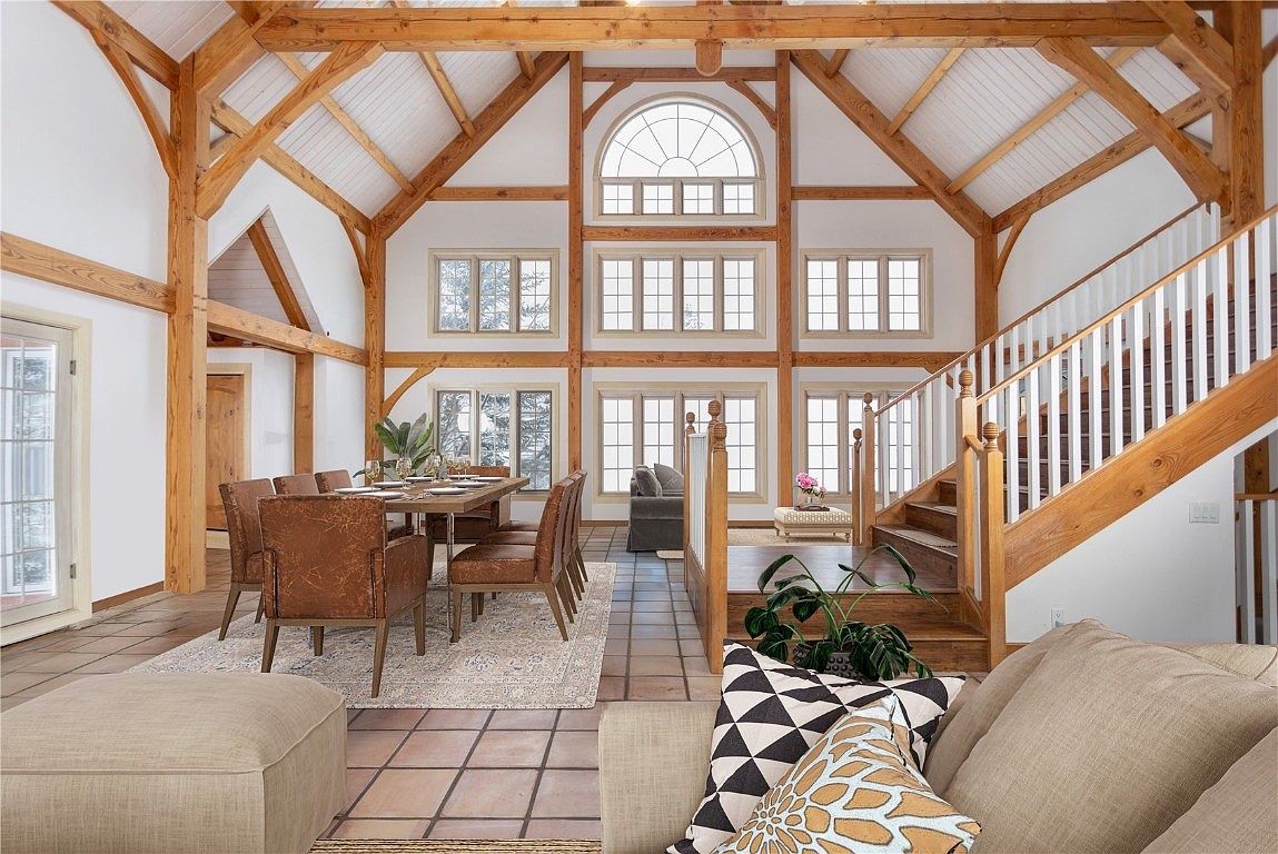 This is an interior shot of a spacious living room featuring a high vaulted ceiling with exposed wooden beams, creating a rustic yet elegant atmosphere. The room is well-lit by large windows, and the decor includes a dining table with chairs, a staircase, and comfortable seating arrangements, all contributing to a warm and inviting ambiance. The camera is positioned at a medium height, capturing the depth and breadth of the room, showcasing its open-concept design and highlighting the architectural details.