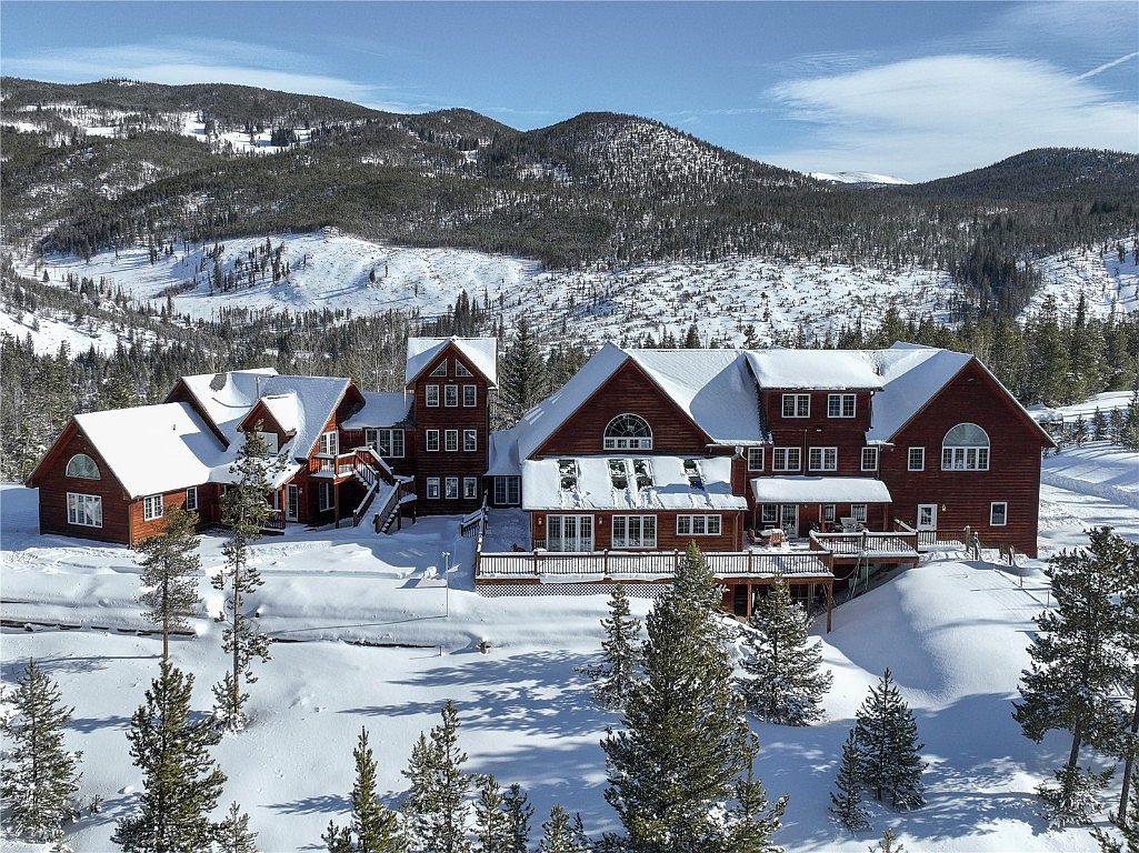 This image showcases a grand, multi-sectioned log home nestled in a snowy, mountainous landscape. The architecture features a combination of gabled roofs, multiple windows, and a large deck, creating a luxurious and inviting atmosphere. The surrounding snow-covered terrain and evergreen trees enhance the property's secluded and picturesque setting.