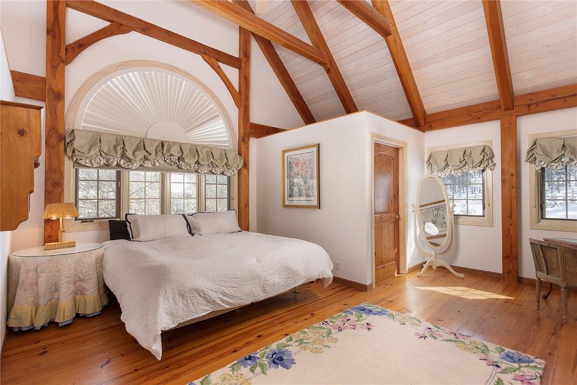 This is a cozy primary bedroom featuring exposed wooden beams and a vaulted ceiling, creating a rustic yet inviting atmosphere. The room is well-lit with natural light streaming through multiple windows, complemented by soft window treatments. A comfortable bed with white linens is the focal point, and a decorative rug adds warmth to the hardwood floors.