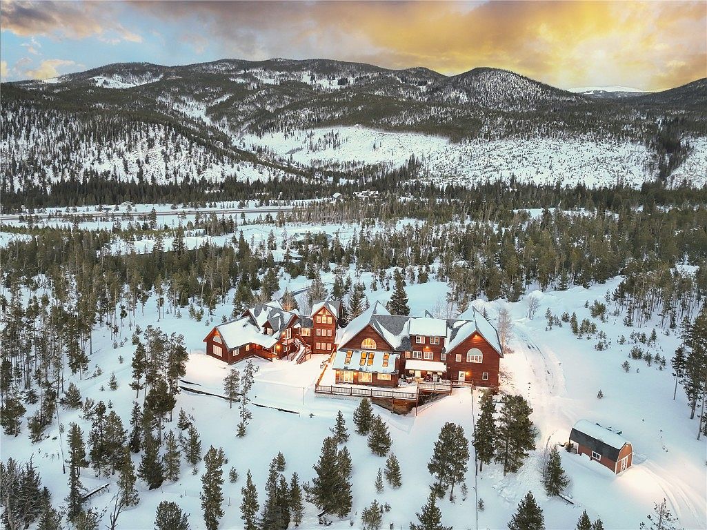 This aerial view showcases a large, luxurious mountain home nestled in a snow-covered landscape. The property features multiple interconnected structures with dark roofs, surrounded by evergreen trees and distant mountain ranges. A long driveway leads to the house, and a separate outbuilding is visible nearby, creating a sense of privacy and seclusion.
