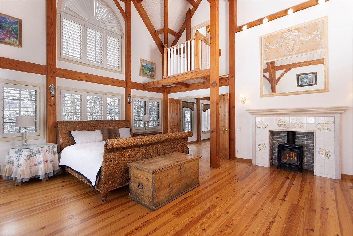This is a spacious primary bedroom featuring exposed wooden beams and a high ceiling, creating a rustic yet elegant atmosphere. The room includes a woven bed frame, a wooden chest, and a fireplace with decorative tiles. Natural light floods the space through large windows, highlighting the hardwood floors and the overall warmth of the room.