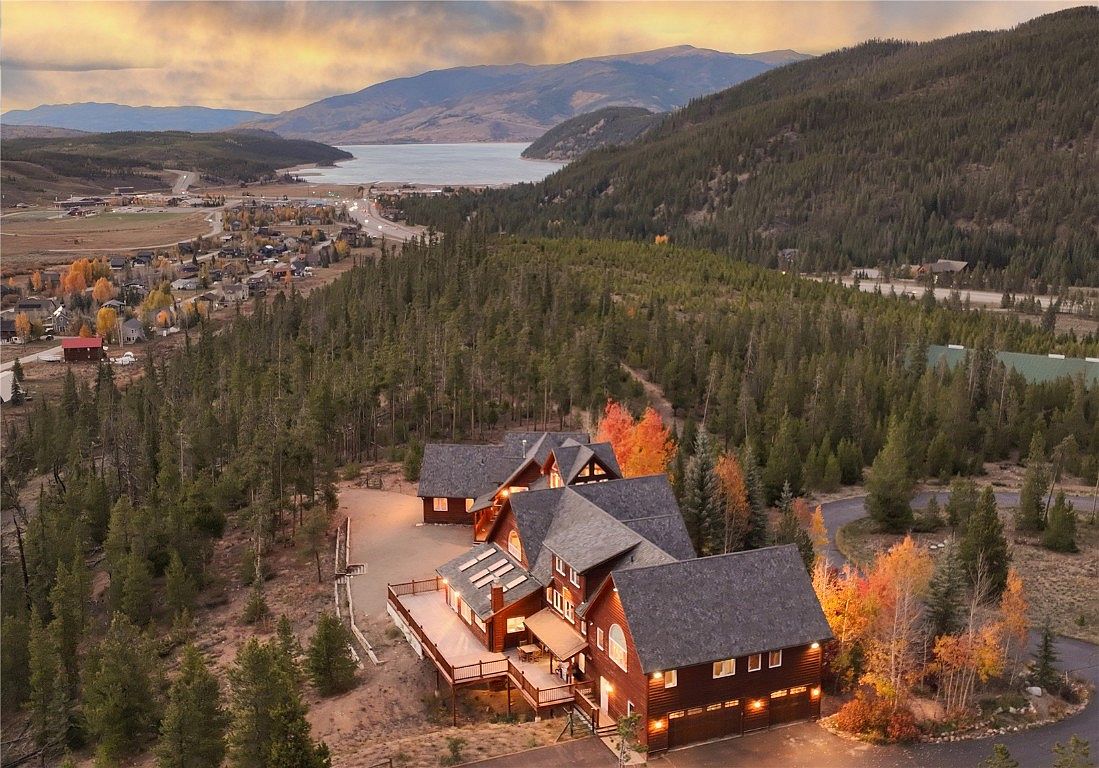 This aerial view showcases a large, luxurious home nestled in a wooded area with a lake and mountains in the background. The house features a complex roofline, multiple decks, and an attached garage. The surrounding landscape includes a mix of evergreen trees and fall foliage, creating a picturesque and serene setting.