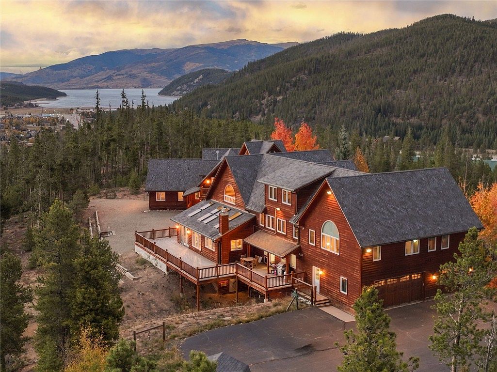 This aerial view showcases a large, multi-story wooden house with a dark gray roof, nestled among tall pine trees. The house features a spacious deck, multiple windows, and an attached garage. In the background, a lake and mountains create a scenic backdrop, enhancing the property's appeal.