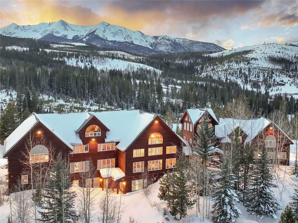 This image showcases the front exterior of a large, luxurious mountain home during winter. The multi-story residence features a dark wood facade, snow-covered roofs, and numerous windows that suggest ample natural light. The property is surrounded by snow-covered ground, evergreen trees, and a stunning mountain backdrop, creating a serene and picturesque setting.