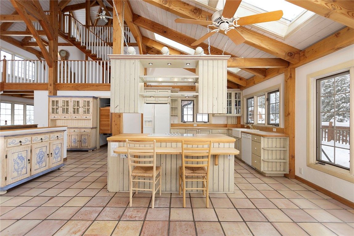 This is a spacious kitchen with a rustic, farmhouse-style design. It features exposed wooden beams, a central island with seating, and custom cabinetry with a distressed finish. The kitchen also includes modern appliances and large windows that provide natural light and views of the snowy outdoors.