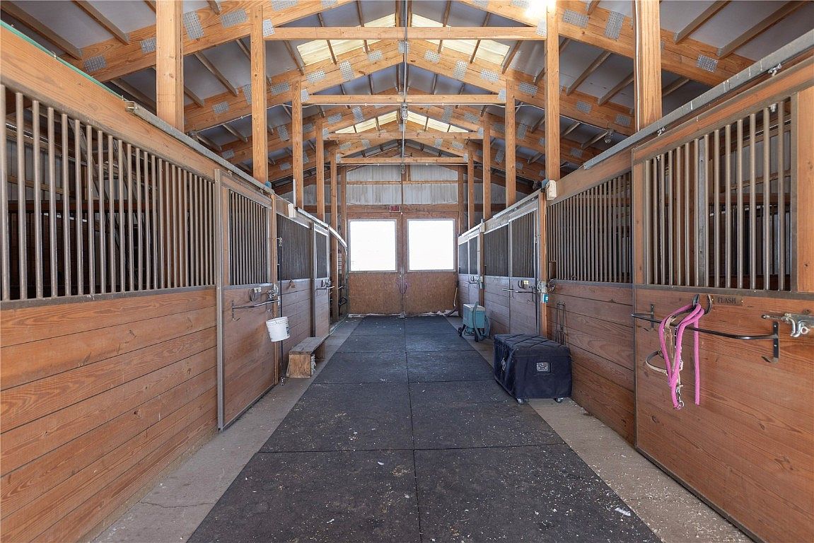 This image showcases the interior of a horse barn, featuring wooden stalls with metal bars on either side of a central aisle. The aisle is covered with black rubber mats, and natural light streams in through windows at the end of the barn and skylights in the roof. The perspective is a straight-on view down the center of the aisle, emphasizing the length and structure of the barn.