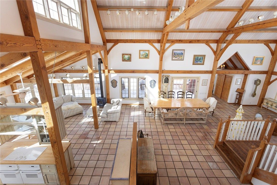 This expansive living room features a soaring ceiling with exposed wooden beams, creating a rustic yet elegant atmosphere. The room is filled with natural light from numerous windows, highlighting the tiled flooring and comfortable seating arrangements. A wood-burning stove adds a cozy touch, while the open layout seamlessly connects the living area with the dining space.