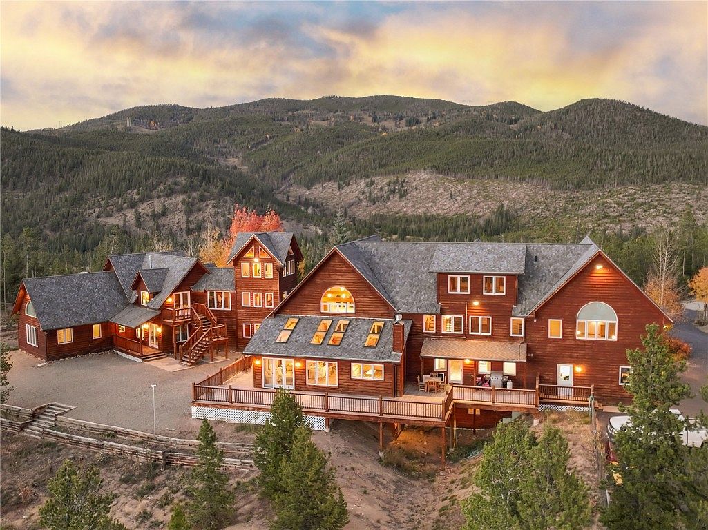 This aerial view showcases a large, multi-structured log home nestled in a mountainous, wooded landscape. The property features multiple gabled roofs, a spacious deck, and a rustic fence, blending seamlessly with the natural surroundings. The overall impression is one of secluded luxury and natural beauty.